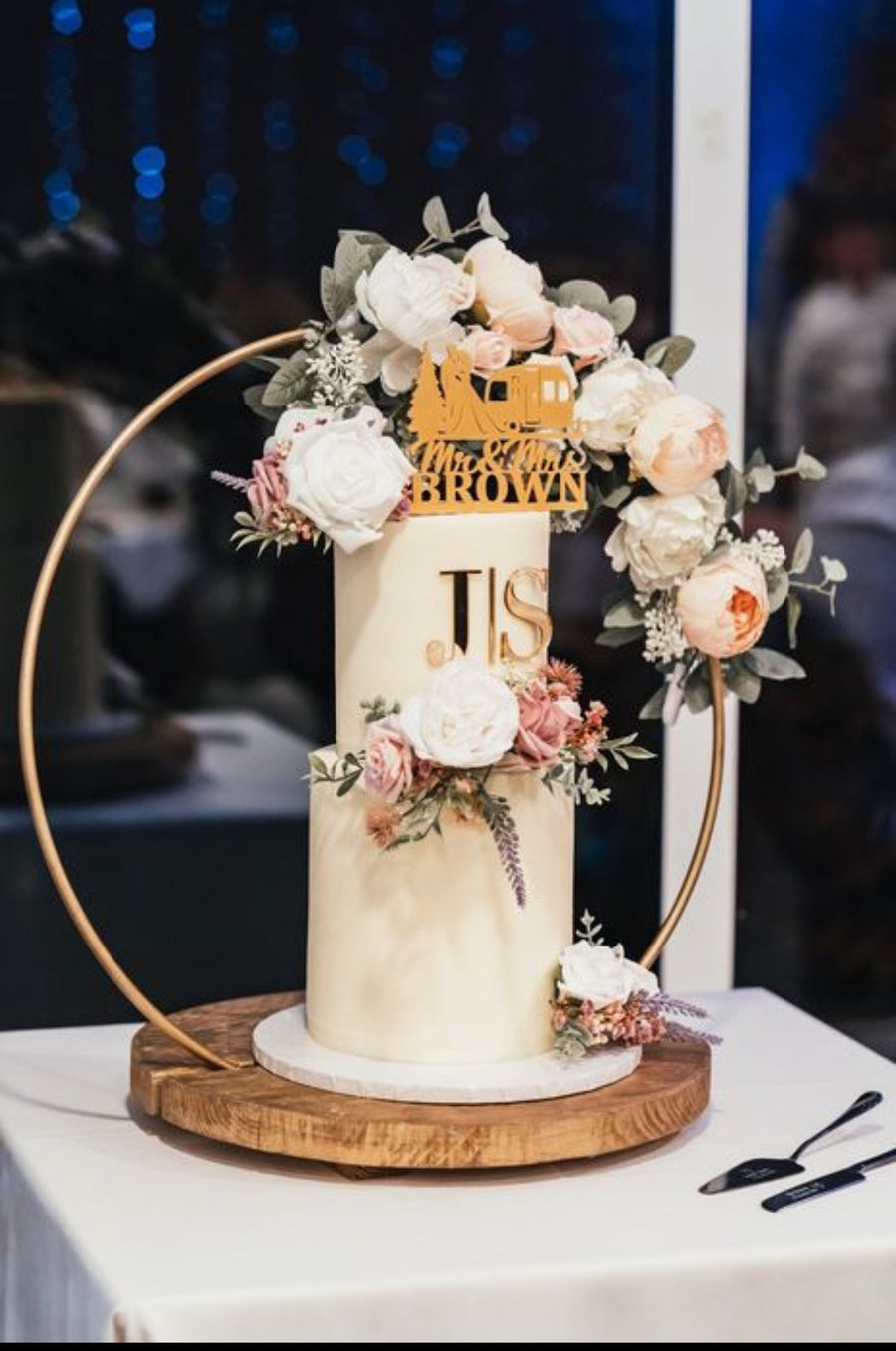 A wedding cake decorated with buttercream and floral hoop stand. The top tier has a personalised topper and a gold acrylic monogram
