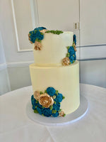 A two-tier wedding cake with buttercream flowers in blue and white colors, placed on a table with a vase of white roses beside it.