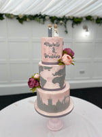 A three-tier fondant covered wedding cake with travel-themed decorations, such as a map and travel suitcase details, and fresh flower decorations.