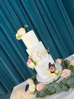 A three-tier wedding cake with white fondant icing, decorated with a monogram and surrounded by fresh flowers on a silver platter.