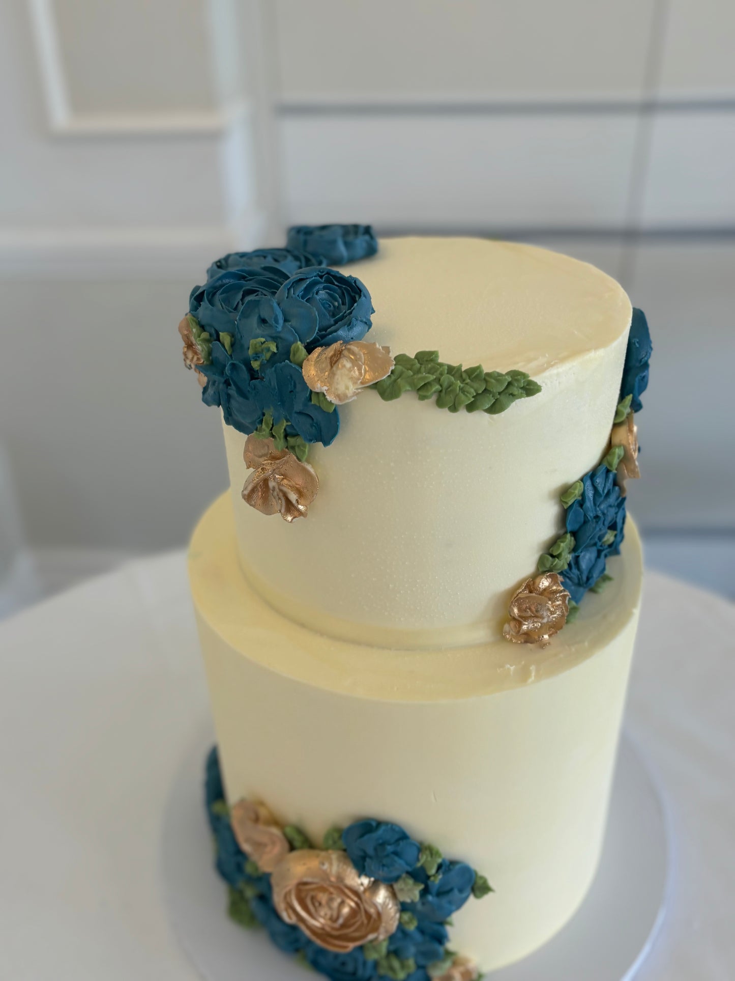 A two-tier wedding cake with buttercream flowers in blue and white colors, placed on a table with a vase of white roses beside it.