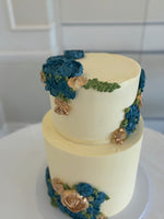 A two-tier wedding cake with buttercream flowers in blue and white colors, placed on a table with a vase of white roses beside it.