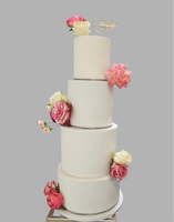A four-tier white fondant wedding cake decorated with pink and white sugar flowers and a golden cake topper at the top.