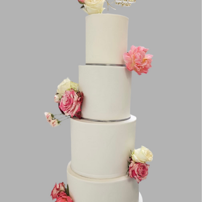 A four-tier white fondant wedding cake decorated with pink and white sugar flowers and a golden cake topper at the top.