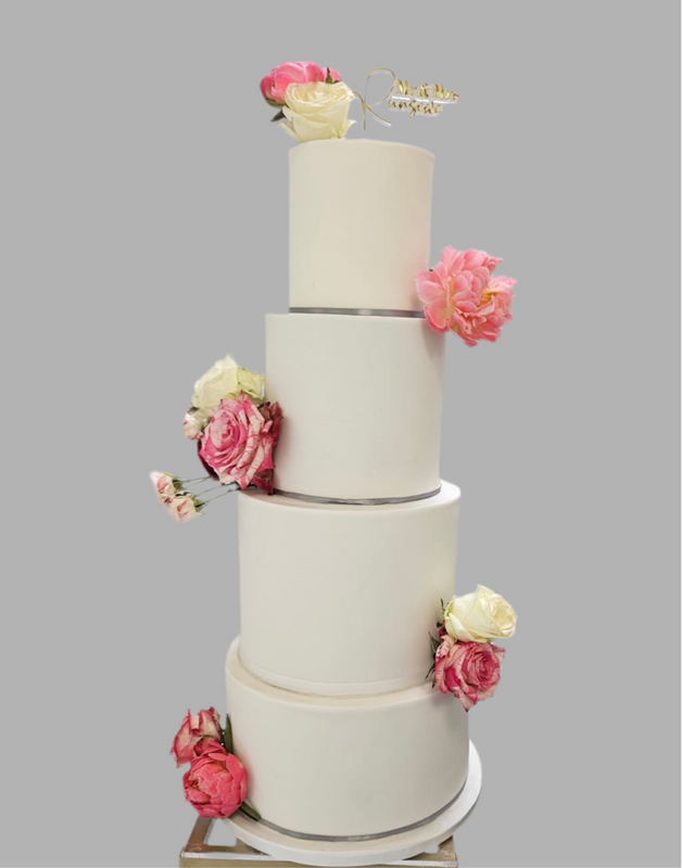 A four-tier white fondant wedding cake decorated with pink and white sugar flowers and a golden cake topper at the top.