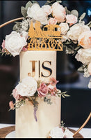 A wedding cake decorated with buttercream and floral hoop stand. The top tier has a personalised topper and a gold acrylic monogram