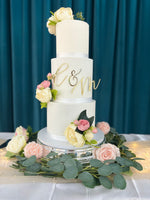 A three-tier wedding cake with white fondant icing, decorated with a monogram and surrounded by fresh flowers on a silver platter.