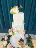 A three-tier wedding cake with white fondant icing, decorated with a monogram and surrounded by fresh flowers on a silver platter.