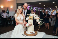 A wedding cake decorated with buttercream and floral hoop stand, being cut by a man in a suit and a woman in a white wedding dress.