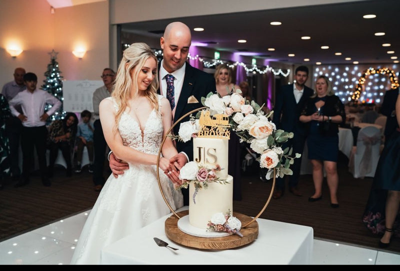 A wedding cake decorated with buttercream and floral hoop stand, being cut by a man in a suit and a woman in a white wedding dress.