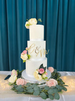 A three-tier wedding cake with white fondant icing, decorated with a monogram and surrounded by fresh flowers on a silver platter.