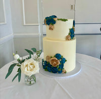 A two-tier wedding cake with buttercream flowers in blue and white colors, placed on a table with a vase of white roses beside it.