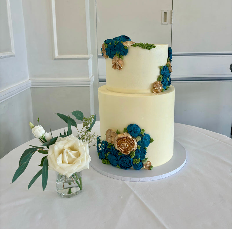 A two-tier wedding cake with buttercream flowers in blue and white colors, placed on a table with a vase of white roses beside it.