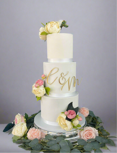 A three-tier wedding cake with white fondant icing, decorated with a monogram and surrounded by fresh flowers on a silver platter.