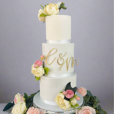 A three-tier wedding cake with white fondant icing, decorated with a monogram and surrounded by fresh flowers on a silver platter.