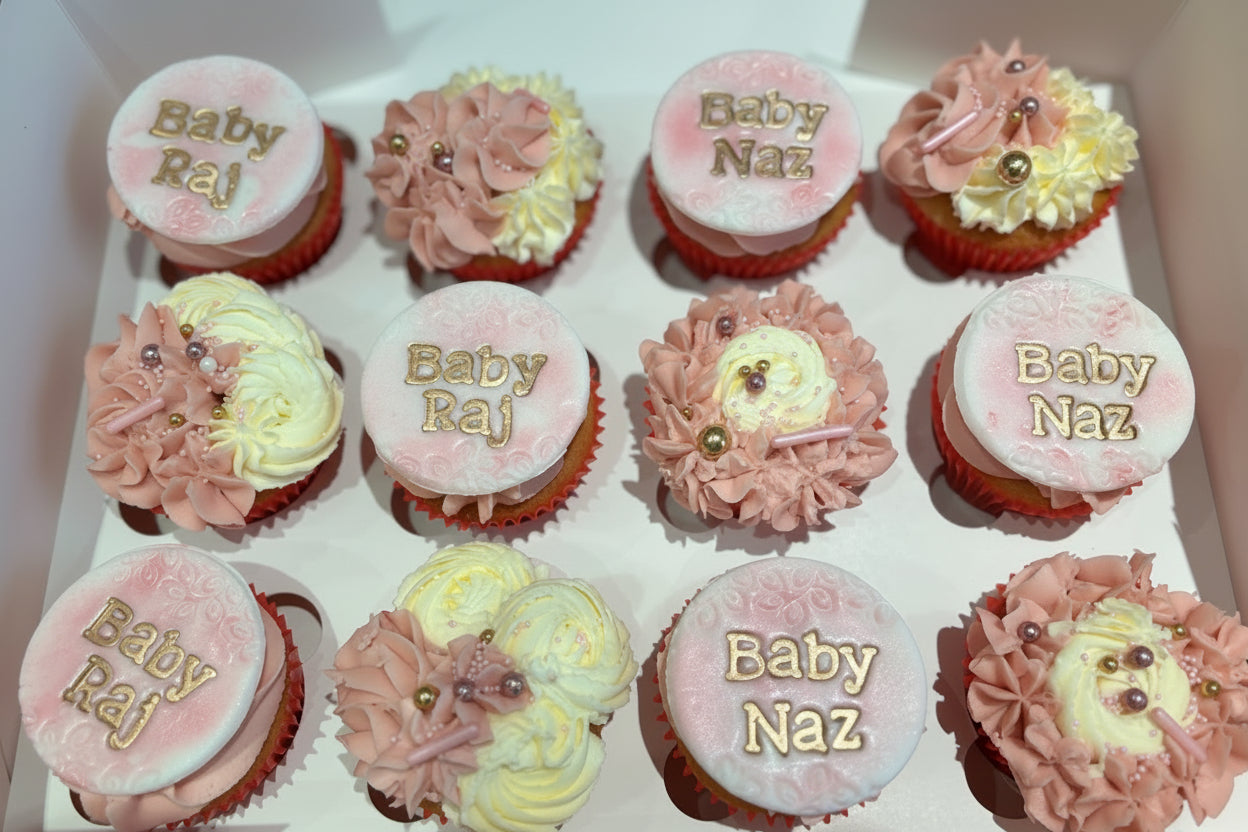 A box of personalised cupcakes for a baby shower, with pink frosting and decorative elements on each cupcake.