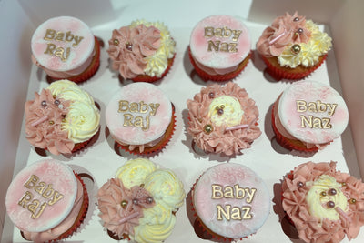 A box of personalised cupcakes for a baby shower, with pink frosting and decorative elements on each cupcake.