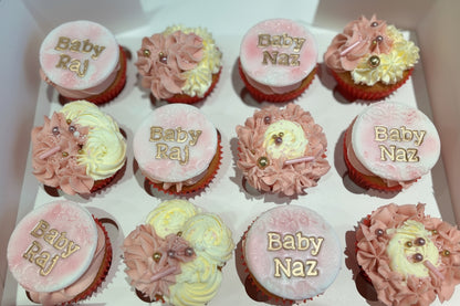 A box of personalised cupcakes for a baby shower, with pink frosting and decorative elements on each cupcake.