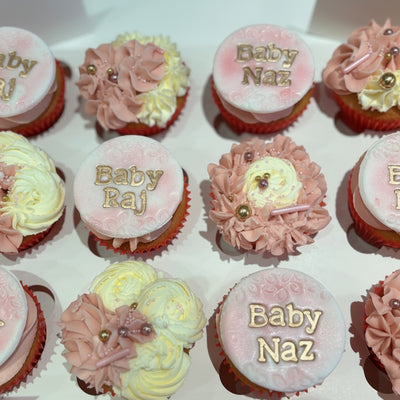 A box of personalised cupcakes for a baby shower, with pink frosting and decorative elements on each cupcake.