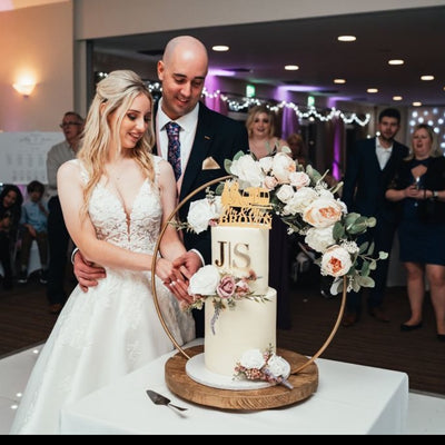 A wedding cake decorated with buttercream and floral hoop stand, being cut by a man in a suit and a woman in a white wedding dress.