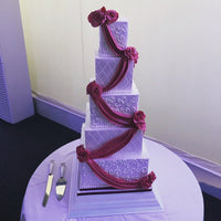 A five-tier square wedding cake with white fondant icing and dark red decorative ribbons, displayed on a table with a cake server and knife.