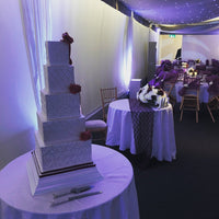 A five-tier square wedding cake with white fondant icing and dark red decorative ribbons, displayed on a table with a cake server and knife.