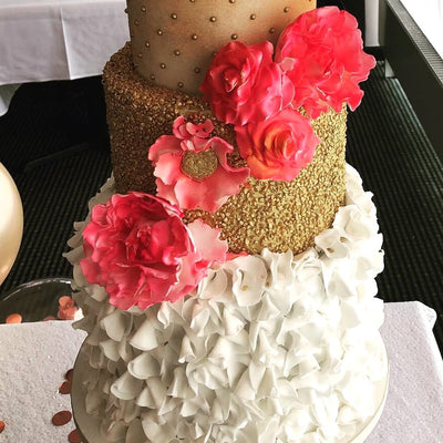 A three-tiered wedding cake with a combination of white fondant ruffles and golden tiers, decorated with pink sugar flowers.