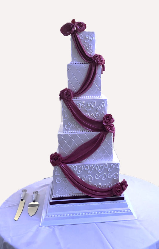 A five-tier square wedding cake with white fondant icing and dark red decorative ribbons, displayed on a table with a cake server and knife.