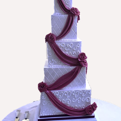A five-tier square wedding cake with white fondant icing and dark red decorative ribbons, displayed on a table with a cake server and knife.