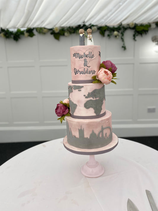 A three-tier fondant covered wedding cake with travel-themed decorations, such as a map and travel suitcase details, and fresh flower decorations.