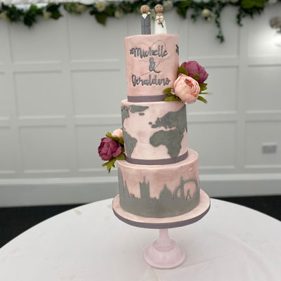 A three-tier fondant covered wedding cake with travel-themed decorations, such as a map and travel suitcase details, and fresh flower decorations.