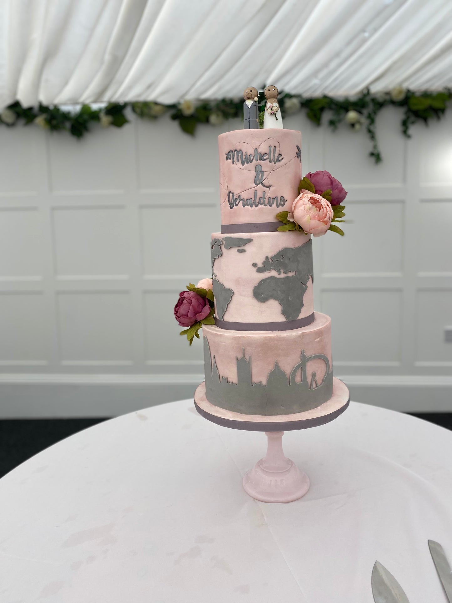A three-tier fondant covered wedding cake with travel-themed decorations, such as a map and travel suitcase details, and fresh flower decorations.